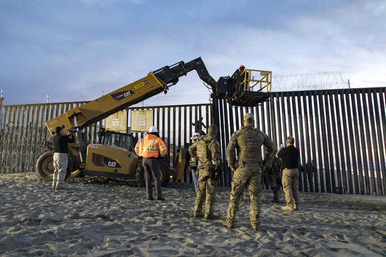 US border patrol stands watch as barbed wire is added to a border wall, 2018.